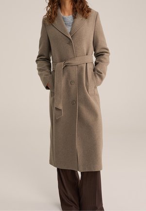 Femme portant un manteau en laine beige mi-long avec ceinture, fermeture à boutons, et les mains dans les poches, portant un pull gris et un pantalon marron.