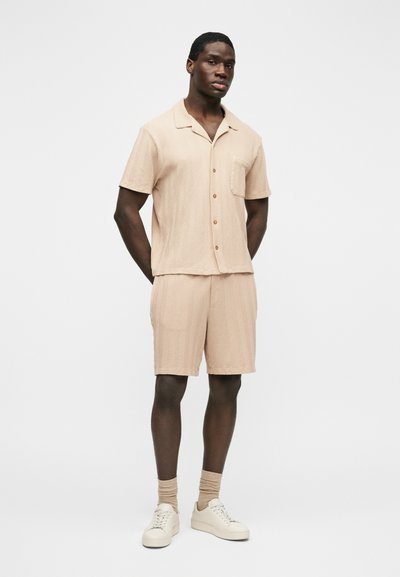 Homme debout, les mains derrière le dos, portant une chemise beige à manches courtes avec boutons, un short assorti, des chaussettes beiges et des baskets blanches sur fond blanc.