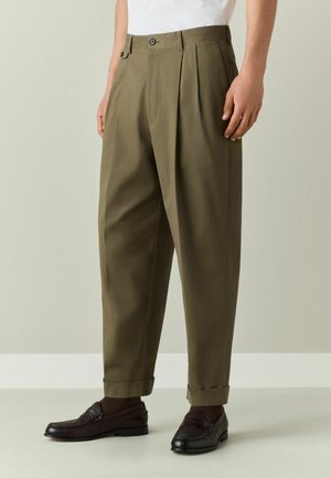 Mann trägt olivgrüne plissierte Hosen mit umgeschlagenen Bündchen an den Knöcheln und dunkelbraune Lederloafer, steht vor einem neutralen Hintergrund.