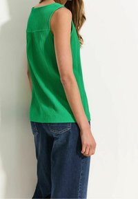 Grön, strukturerad ärmlös topp med en subtil plisserad design, matchad med lösa blå jeans. Toppen har en rund halsringning och en rak fåll.