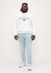 Biely mikina s logom, predným vreckom a sťahovacou šnúrkou. Svetlomodré slim-fit džínsy. Biely tenisky s hnedými akcentmi. Jednoduché pozadie.