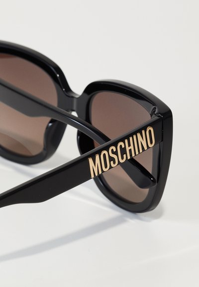 Černé plastové sluneční brýle s hnědými skly. Mají výrazné, obdélníkové obroučky a zlaté logo "Moschino" na boku.