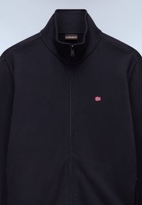 Μαύρο μπουφάν με φερμουάρ και ψηλό γιακά, κατασκευασμένο από λείο ύφασμα. Διαθέτει ένα μικρό κεντημένο νορβηγικό σημαία στην αριστερή πλευρά του στήθους.