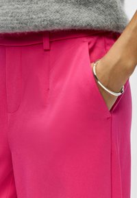 Pantalon rose en tissu lisse, doté de poches latérales et d'une coupe ajustée. Un bracelet argenté orne le poignet, rehaussant le style.
