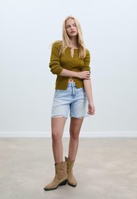 Maslinasto zelena rebrasta cardigan s dugmadi, izlizani svijetloplavi traper šorc i tamne kaubojke s ukrasnim šavovima.