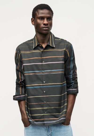 Άνδρας που φοράει σκούρο πουκάμισο με κουμπιά και πολύχρωμες οριζόντιες ρίγες, και ανοιχτό μπλε τζιν, στέκεται με τα χέρια στις τσέπες.