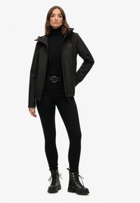 Black waterproof jacket with a hood, featuring a logo on the chest, paired with a black turtleneck and fitted pants, worn with black combat boots.