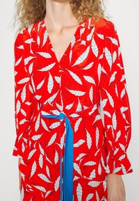 Rotes Kleid mit weißen Blattmustern, V-Ausschnitt, langen Ärmeln und einem blauen Stoffgürtel, der in der Taille gebunden ist. Glattes, leichtes Material.