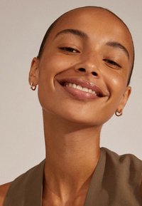 Naeratav inimene sujuva jume ja kuldsete rõngastega kõrvarõngastega. Taust on neutraalne. Riietus on valmistatud pehmest oliivrohelisest kangast.