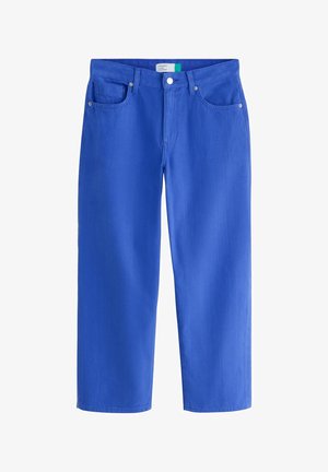 Een vrouw staat met haar handen in haar zakken, draagt een wit asymmetrisch tanktop, blauwe wijde broekspijpen en blauw-witte sneakers.