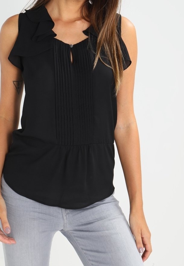 Femme portant un chemisier noir sans manches avec un col froufroutant et des plis, associé à un jean gris clair sur fond uni.