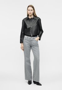 Žena stojaca v čiernej koženej bunde na zips, svetlosivých širokých vysokopásových džínsach a čiernych čižmách so špicatou špičkou, pózujúca na bielom pozadí.