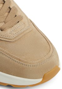Zapatilla de ante beige con costuras prominentes, punta redondeada y superficie texturizada. Presenta una suela blanca y una suela de goma con detalles en marrón claro.