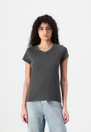 Jeune femme aux cheveux bouclés et foncés, portant un t-shirt ajusté gris foncé à manches courtes et un jean bleu clair, debout sur fond blanc.