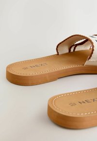 Paire de sandales plates couleur beige avec des brides en tissu blanc et une bordure marron, affichant le nom de la marque "NEXT" et la taille "5 UK 38 EUR" sur les semelles.