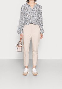 Donna che indossa pantaloni beige a vita alta, blusa a maniche lunghe floreale, sneakers bianche e beige, mentre tiene una borsa beige con tracolla marrone.