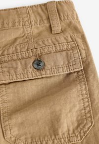 Tan stofshorts med et struktureret mønster, en baglomme med en mørk knap og forstærket syninger langs sømmene.