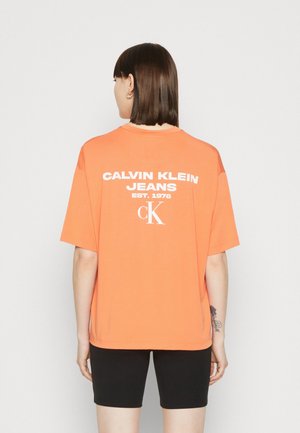 Persoon met schouderlang bruin haar draagt een losse oranje Calvin Klein Jeans T-shirt met witte logo tekst, gecombineerd met een zwarte short, staand met de rug naar de kijker.