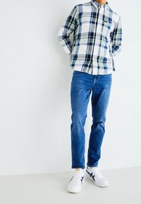 Camisa de cuadros en blanco, verde y marino, con cuello abotonado. Usada con jeans azules y zapatillas blancas con acentos en marino.