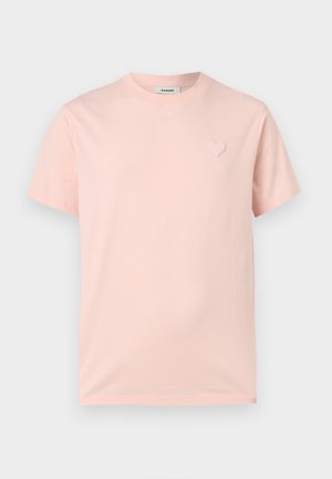 Mand med krøllet hår stående, iført en lyserød t-shirt, sorte bukser og sorte sko mod en ensartet lys baggrund.