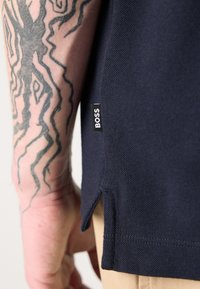 Mornarsko modra polo majica z teksturirano površino, z bočnim razporkom in majhnim črnim oznako "BOSS" na robu. Odkrit tatoo na roki.