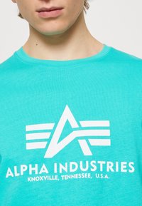 Türkisfarbiges T-Shirt mit kurzen Ärmeln aus Baumwolle, mit einem großen weißen Grafiklogo und dem Text "ALPHA INDUSTRIES" auf der Vorderseite.