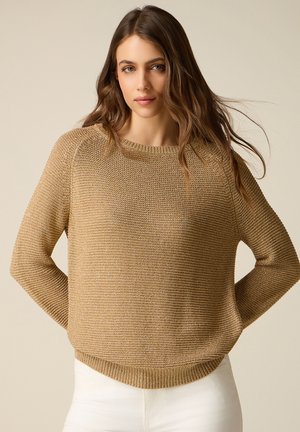 Kvinde med langt brunt hår iført en løs beige striktrøje og hvide bukser, stående mod en ensfarvet lys baggrund.