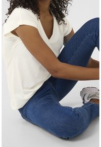 Hvid V-hals T-shirt med oprullede ærmer, parret med blå slim-fit jeans. Model sidder med benene krydset og har sporty hvide sneakers på.