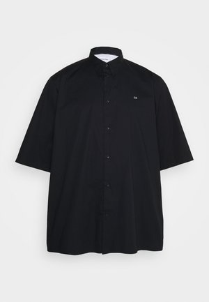 Schwarzes kurzärmliges Hemd mit Knöpfen und Kragen, kleines weißes "CK"-Logo auf der linken Brust, auf hellem einfarbigem Hintergrund dargestellt.