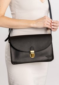 Bolso cruzado de cuero negro con una solapa redondeada, cierre de metal dorado y correa para el hombro. Presenta una textura suave y detalle de costura.