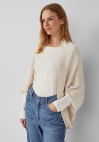 Poncho lavorato a maglia color beige chiaro con punti aperti, design drappeggiato. Indossato sopra una maglietta bianca e abbinato a jeans blu a vita alta.