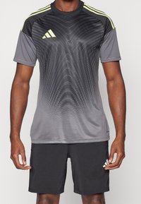 Grå atletisk tröja med ett vågmönster, svarta ärmar och gula detaljer. Matchas med svarta shorts som har en liten logotyp på benet.