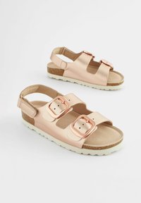 Ett par ljusrosa sandaler med två stora roséguldbuckles och vita texturerade sulor på en enkel vit bakgrund.