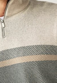 Pull-over beige a maglia con colletto con zip, caratterizzato da un design texturizzato e righe orizzontali in verde scuro e beige. Tessuto morbido, vestibilità casual.