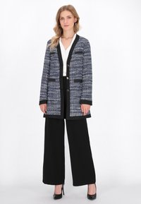 Giacca a fantasia blu navy e bianca con rifiniture e tasche nere, abbinata a pantaloni larghi neri e tacchi neri, il tutto sopra una maglietta bianca.