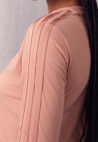 Light pink long-sleeve top with ribbed neck and three vertical stripes along the sleeves, made of a soft, stretchy fabric.