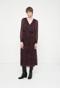 Burgundy pitkähihaista mekkoa, jossa on V-pääntie, kiristetty vyötärö ja virtaava kangas, jossa on aaltoilevia mustia viivoja ja kerrostettua rakennetta. Mustat nilkkurit.