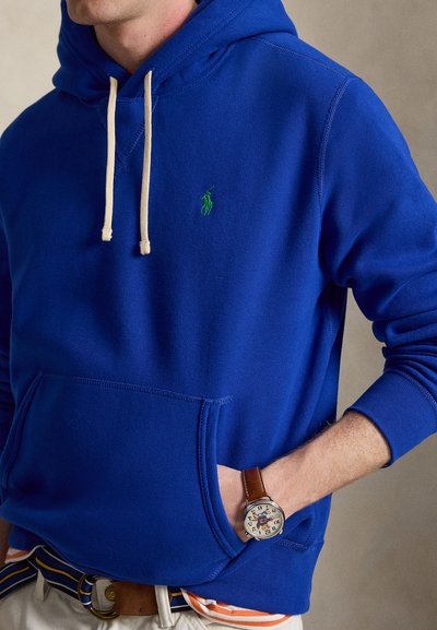 Homme portant un sweat à capuche bleu vif avec des cordons blancs, un logo brodé vert et une montre avec un bracelet en cuir marron au poignet.