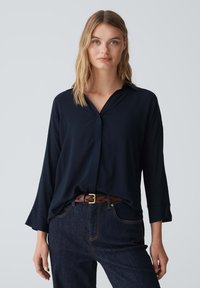 Blusa blu navy a maniche lunghe con colletto, caratterizzata da una vestibilità ampia e un leggero lucido. Abbinata a jeans scuri e una cintura intrecciata marrone.