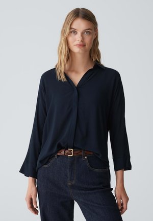 Navy blue, long-sleeve blouse with a collar, featuring a loose fit and a subtle sheen. Paired with dark jeans and a braided brown belt.