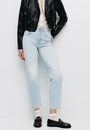 Jean en denim bleu clair à coupe droite, taille haute et finition délavée, associé à une veste en cuir noire et des mocassins sombres brillants.