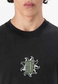 Černé tričko s žebrovaným kulatým výstřihem; má vyšívaný zeleno-bílý design slunečního záření se slovem "HUF" uprostřed.
