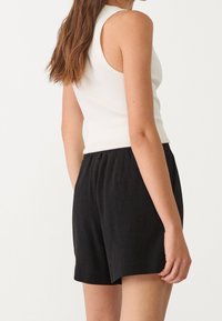 Femme avec de longs cheveux bruns, portant un haut sans manches blanc à côtes et un short noir ample, se tenant devant un fond blanc uni.