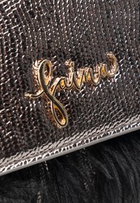 Sort tekstureret stofpose med metallisk finish, der har et guld skrifttype-logo i kursiv. Bemærkelsesværdig fjeragtig detalje nederst.