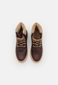 Botas de tornozelo em couro castanho com colarinho de camurça bege, atacadores bege, bico arredondado e sola de borracha. Apresenta dois ilhós de metal de cada lado.