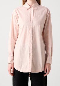 Kvinna iklädd en ljusrosa långärmad skjorta med krage, i kombination med svarta byxor med subtila utsmyckningar.