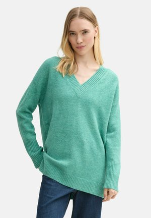 Grüner V-Ausschnitt-Pullover aus einem weichen, strukturierten Stoff mit gerippten Bündchen und Saum, kombiniert mit dunklen Jeans. Lockere, entspannte Passform.