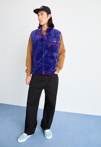 Purple fleece vest with red accents, front zipper pockets; brown long-sleeve shirt underneath, black pants, and light blue sneakers.