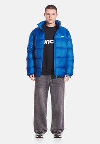 Niebieska kurtka puchowa z wysokim kołnierzem, zapięciem na zamek i ściągaczami przy rękawach. Noszona na czarnym T-shircie z białym nadrukiem, zestawiona z szarymi dżinsami.