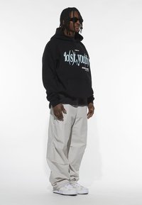 Fekete kapucnis pulóver világoskék grafikus szöveggel, laza világosszürke cargo nadrággal és fehér sportcipővel párosítva. Simább anyag, laza szabás.